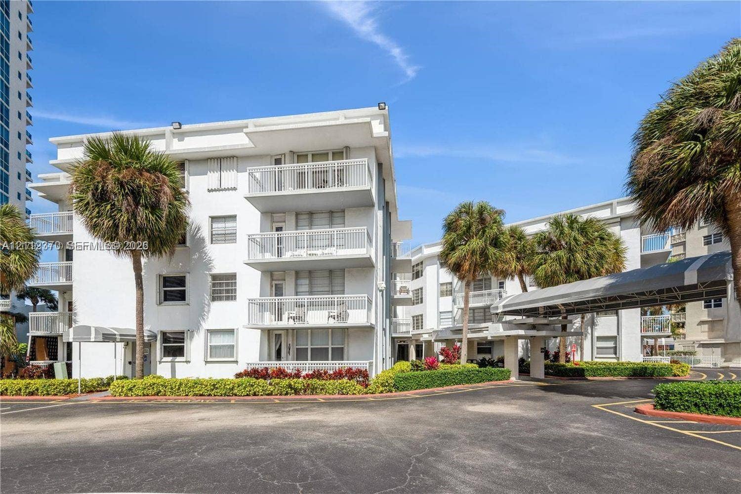 Mid-rise building exterior featuring private balconies with white railings, a covered porte-cochre entry, and mature palm tree landscaping