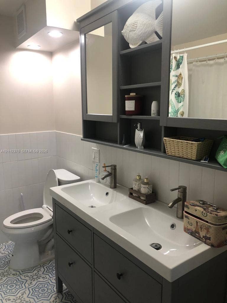 Double vanity featuring a gray cabinet base, integrated dual-basin sink, patterned tile flooring, wall-mounted medicine cabinet with open shelving, and metallic faucets