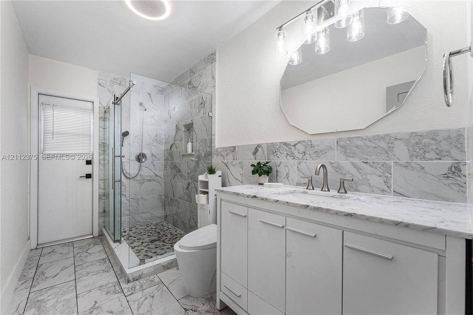 10936 NW 41st Drive, Coral Springs, Coral Springs, FL 33065 Bathroom featuring a white vanity with a stone countertop, an expansive walk-in shower with stone tile surround and pebble flooring, and a large octagonal wall mirror