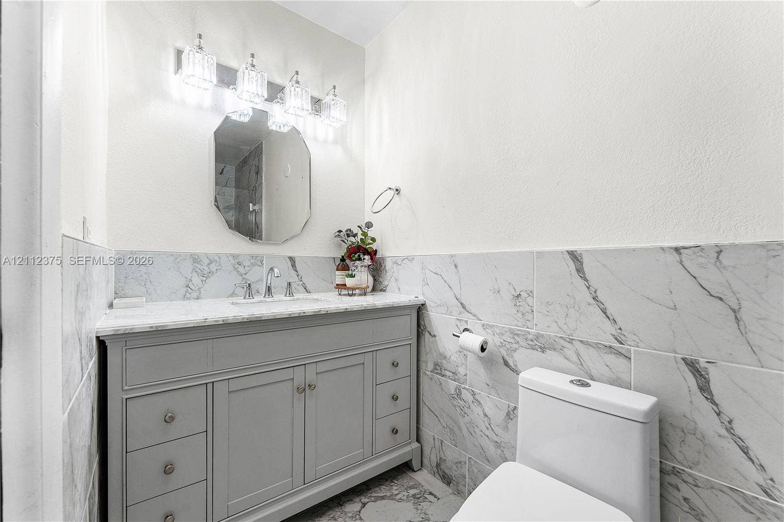 10936 NW 41st Drive, Coral Springs, Coral Springs, FL 33065 Grey vanity with stone-surface countertop, porcelain wall tile with integrated veining, octagonal wall mirror, and four-bulb polished vanity lighting