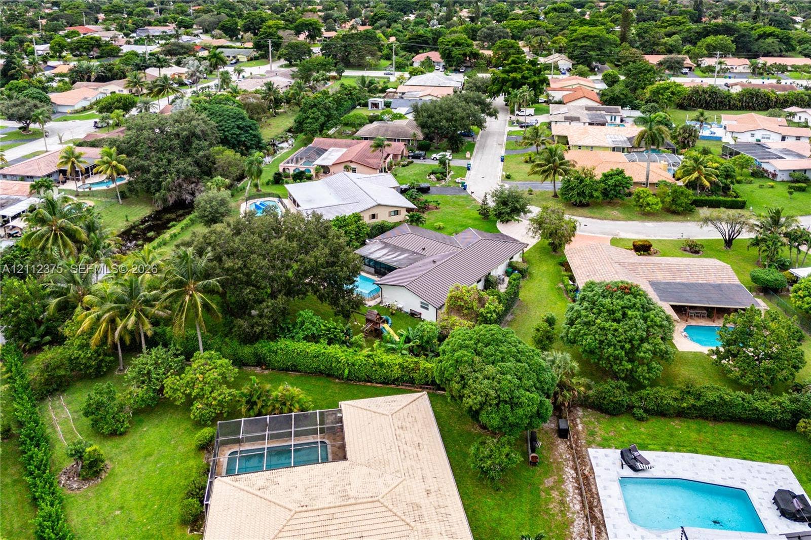 10936 NW 41st Drive, Coral Springs, Coral Springs, FL 33065 Aerial perspective of a residential property featuring an in-ground swimming pool with a screened enclosure, expansive green space, mature landscaping, and a concrete driveway