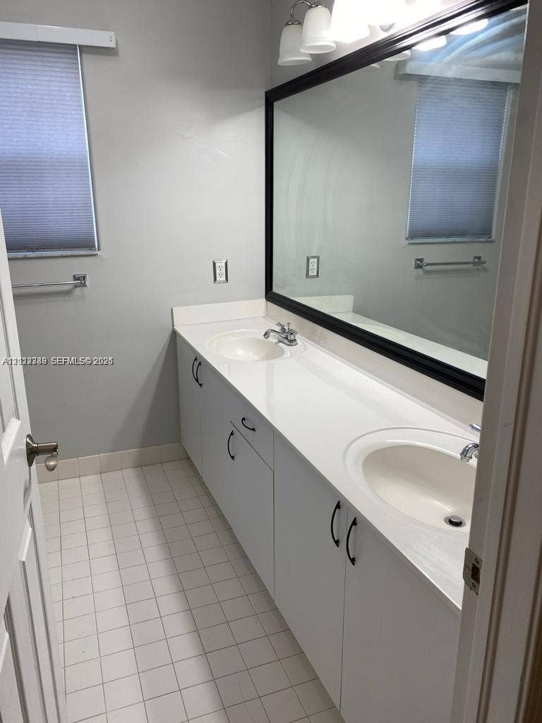 Double vanity with integrated sinks, white countertops, white cabinetry with matte black hardware, large-format mirror with black frame, and tiled flooring