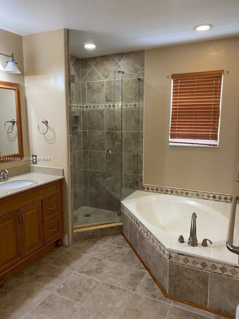 Bathroom featuring a wooden vanity, soaking tub, glass-enclosed shower, stone-finish tile flooring, and decorative tile accents