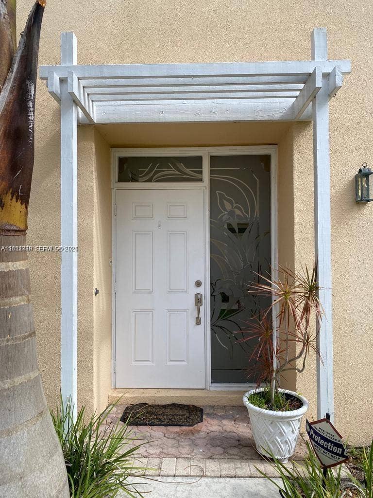 Entrance featuring a white wooden pergola, a paneled door with an etched glass sidelight, a transom window, and stone-paver hardscaping
