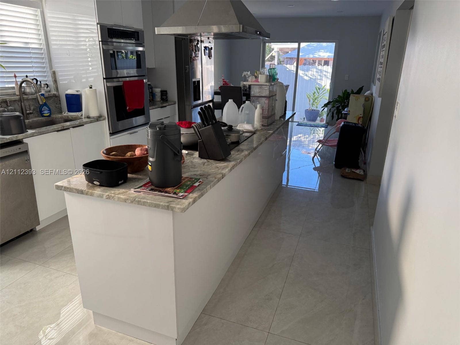 Kitchen featuring a central island with a stone countertop, built-in double ovens, stainless steel range hood, and large-format tile flooring