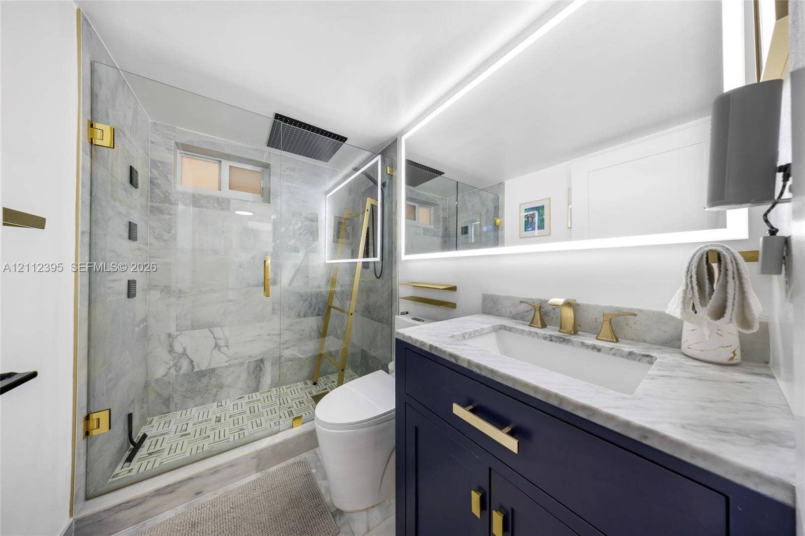 Modern vanity with navy cabinetry, stone countertop, and brass hardware, featuring a glass-enclosed shower with floor-to-ceiling stone tiling, overhead rainfall showerhead, and integrated LED-backlit vanity mirror
