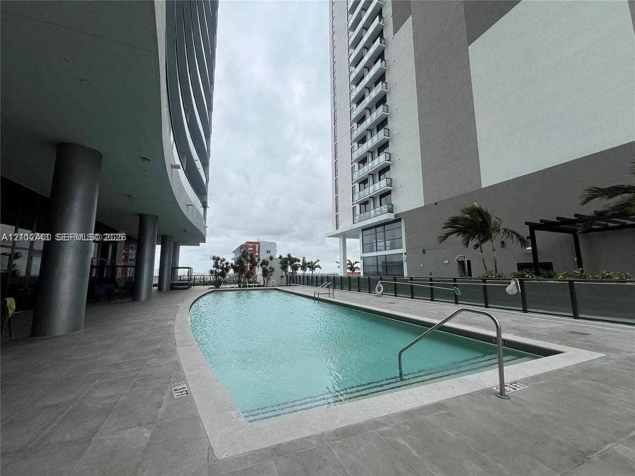 Curved swimming pool featuring a concrete deck, metal handrails, and a glass-panel perimeter fence