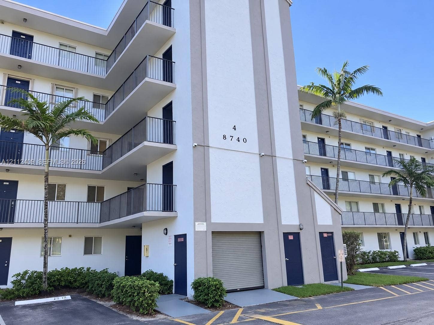 Multi-story building exterior featuring stacked private balconies, metal railings, a ground-level roll-up security door, and exterior landscaping
