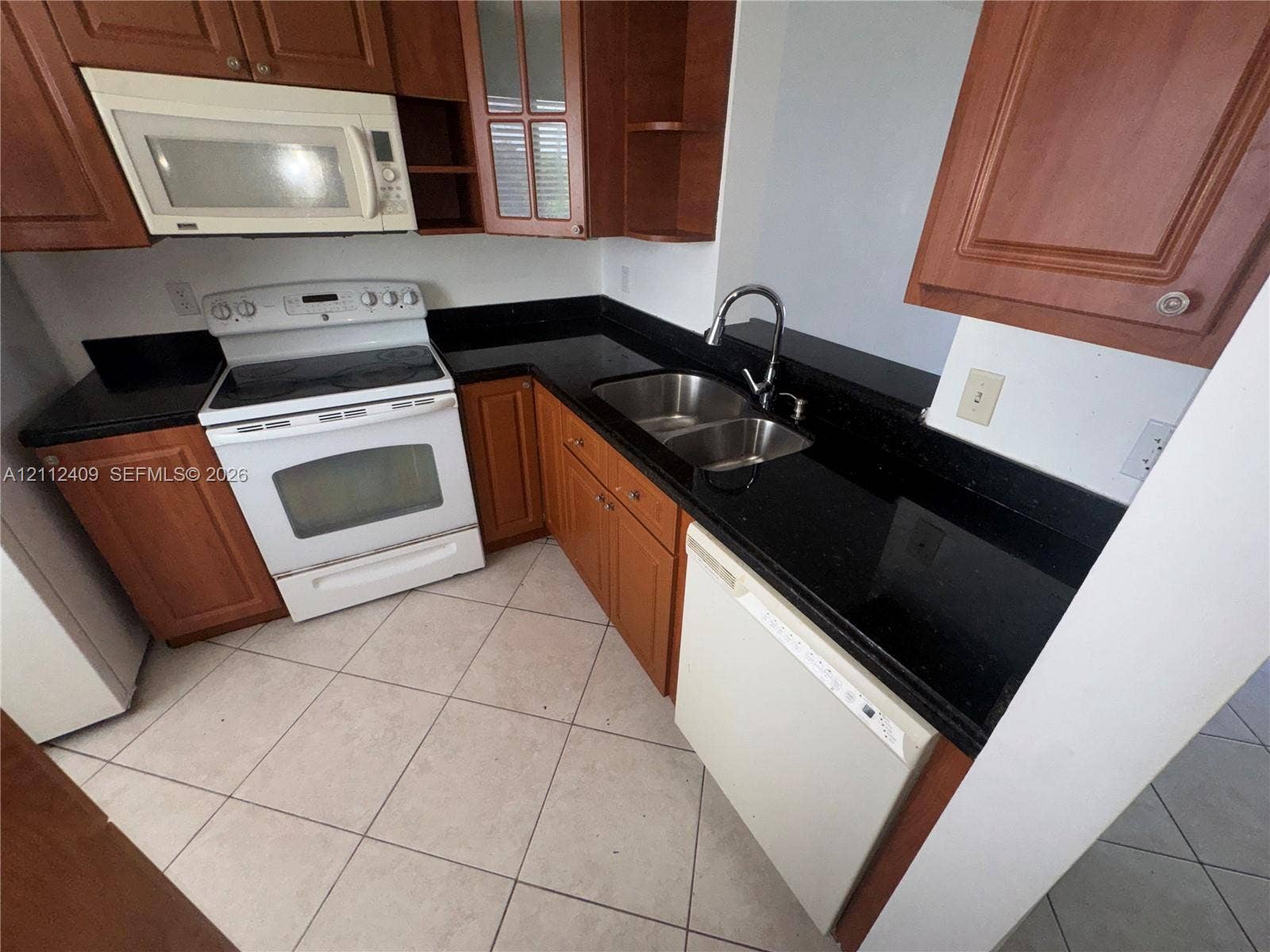 Kitchen featuring wood-finish cabinetry, dark solid-surface countertops, dual-basin stainless steel sink with pull-down faucet, and white appliances including a built-in microwave and dishwasher