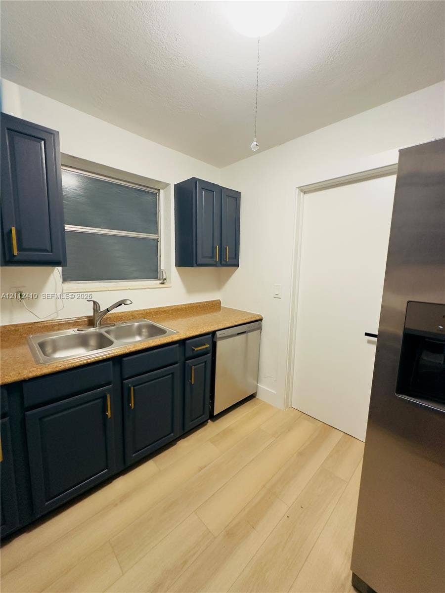 Navy blue cabinetry with gold-toned hardware, stone-finish countertops, stainless steel dual-basin sink, integrated dishwasher, and wood-finish flooring