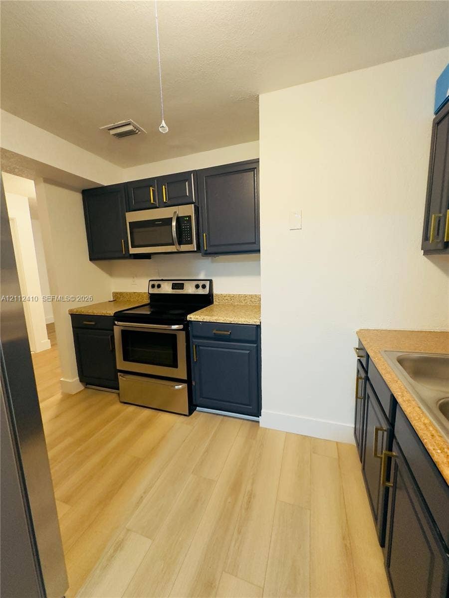 Kitchen featuring navy shaker-style cabinetry with gold-tone hardware, stone-finish countertops, stainless steel appliances, and wood-finish flooring