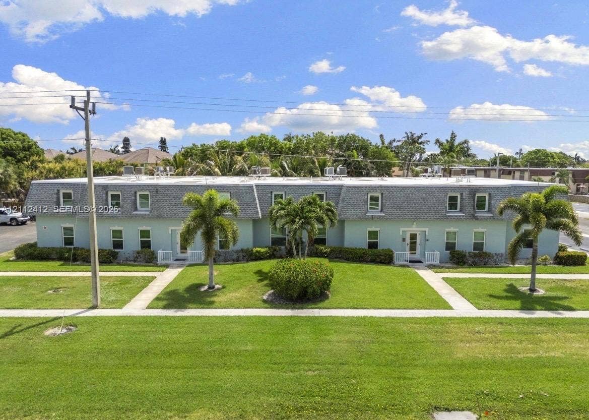 Multi-unit exterior featuring a mansard roofline, light-toned stucco facade, manicured lawn, established palm trees, and concrete walkways