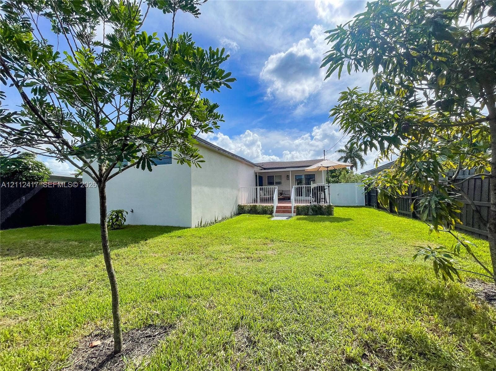 Backyard featuring a manicured lawn, white vinyl fencing, mature trees, and an elevated deck with a white railing