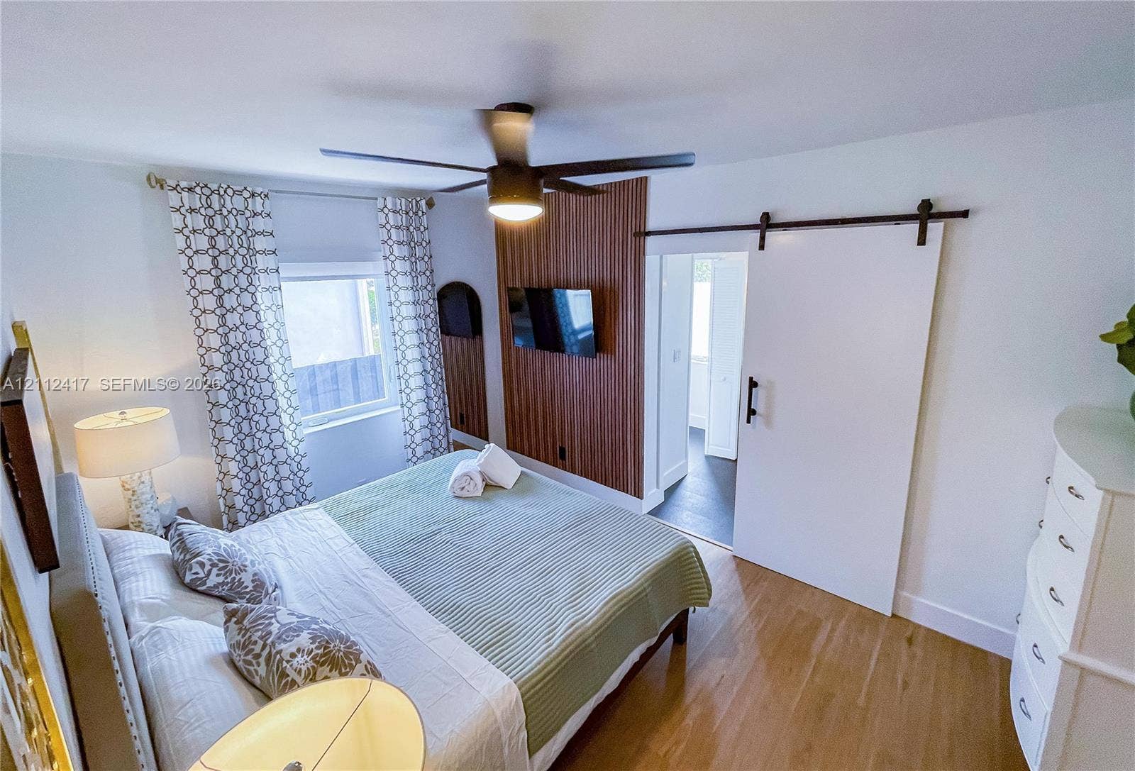 Room featuring wood-finish flooring, a vertical wood-slat accent wall, a white sliding barn door with black hardware, and a contemporary ceiling fan with integrated lighting