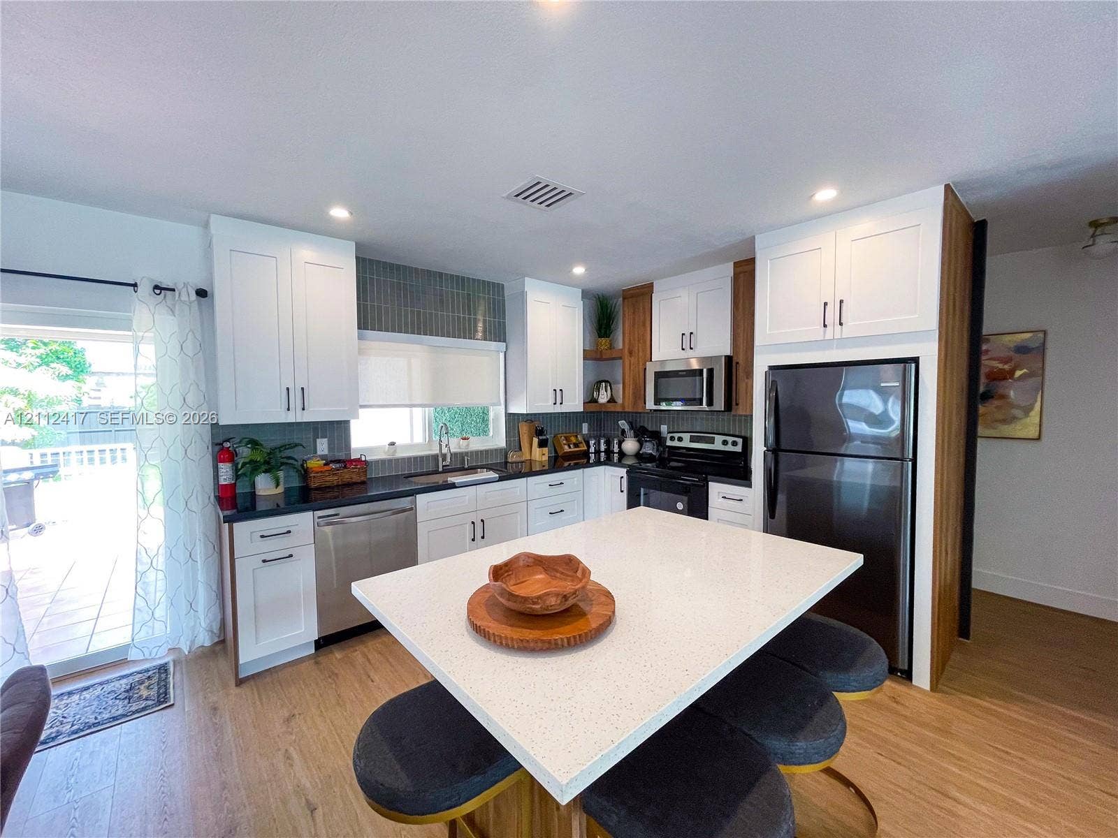 Kitchen featuring white shaker cabinetry, wood-finish flooring, quartz-surfaced central island, stainless steel appliances, and a tiled backsplash
