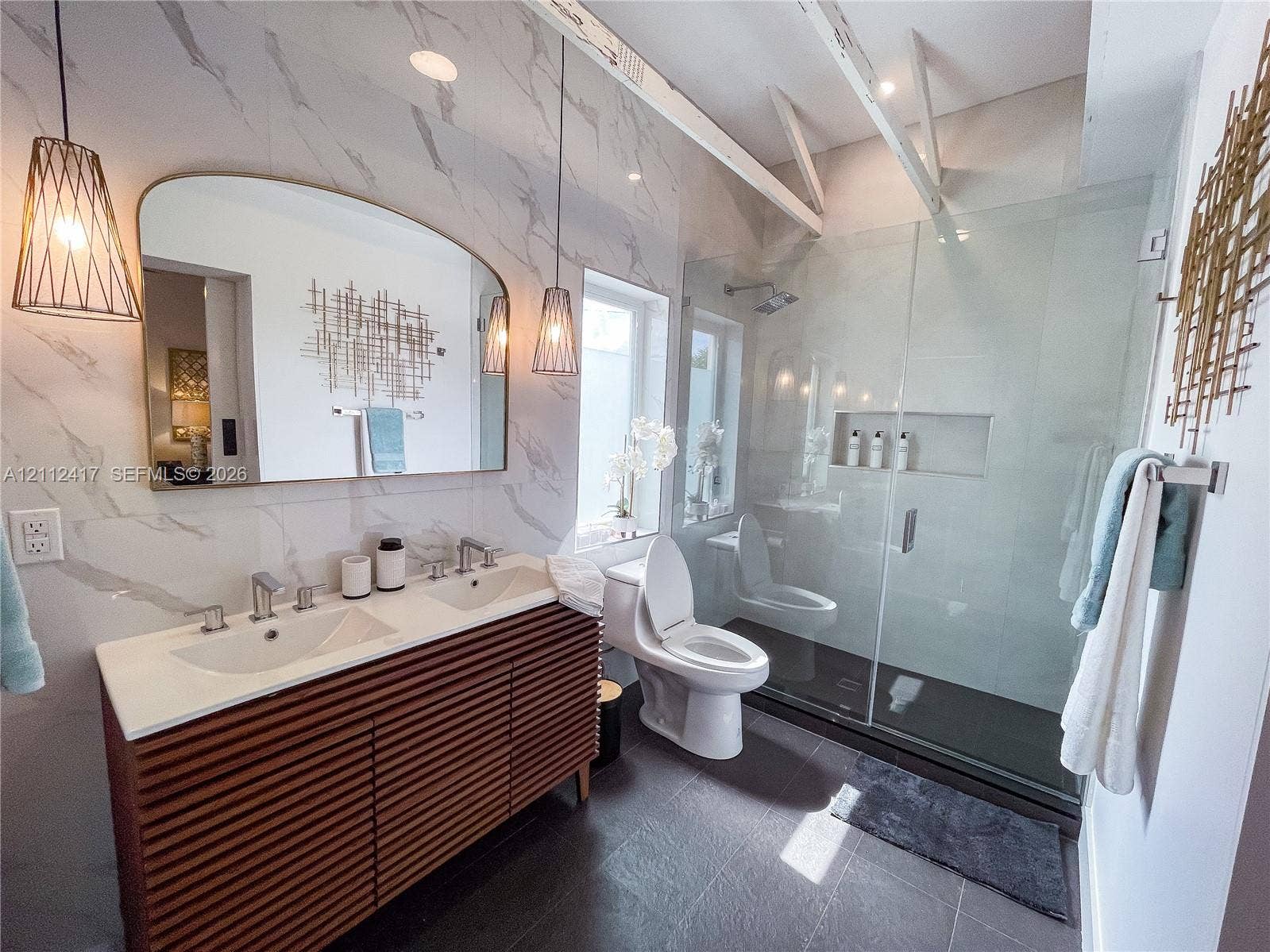 Bathroom featuring a dual-sink vanity with slatted wood cabinet doors, large format stone-finish wall tile, a walk-in glass-enclosed shower with a built-in niche, and dual pendant light fixtures