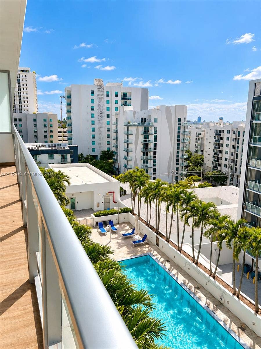 Balcony with wood-finish flooring and metal railing overlooking a swimming pool, palm trees, and an urban landscape