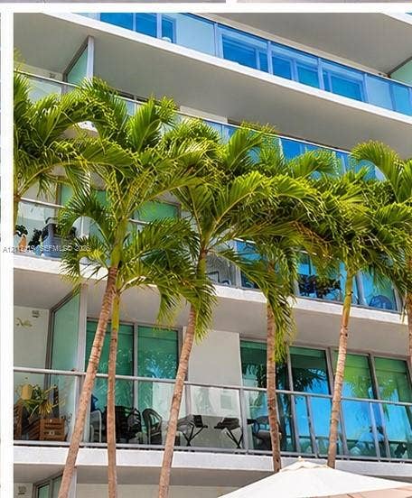 Multi-story exterior featuring floor-to-ceiling glass doors, private balconies with glass railings, and mature palm trees