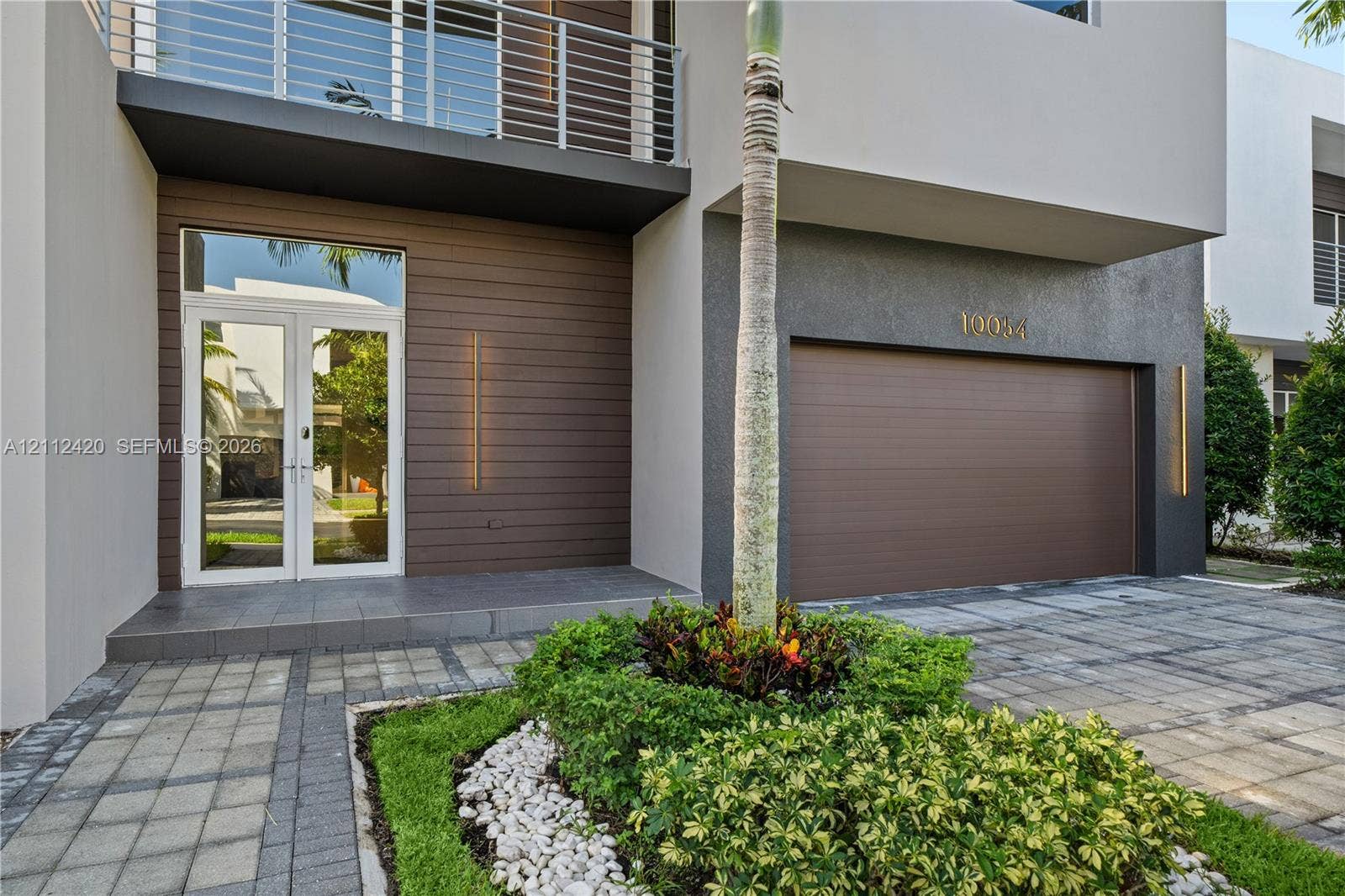 10054 NW 76th Terrace, Doral, FL 33178 Contemporary exterior featuring a horizontal wood-plank facade, second-story balcony with metal railing, double-door entryway, and paver driveway