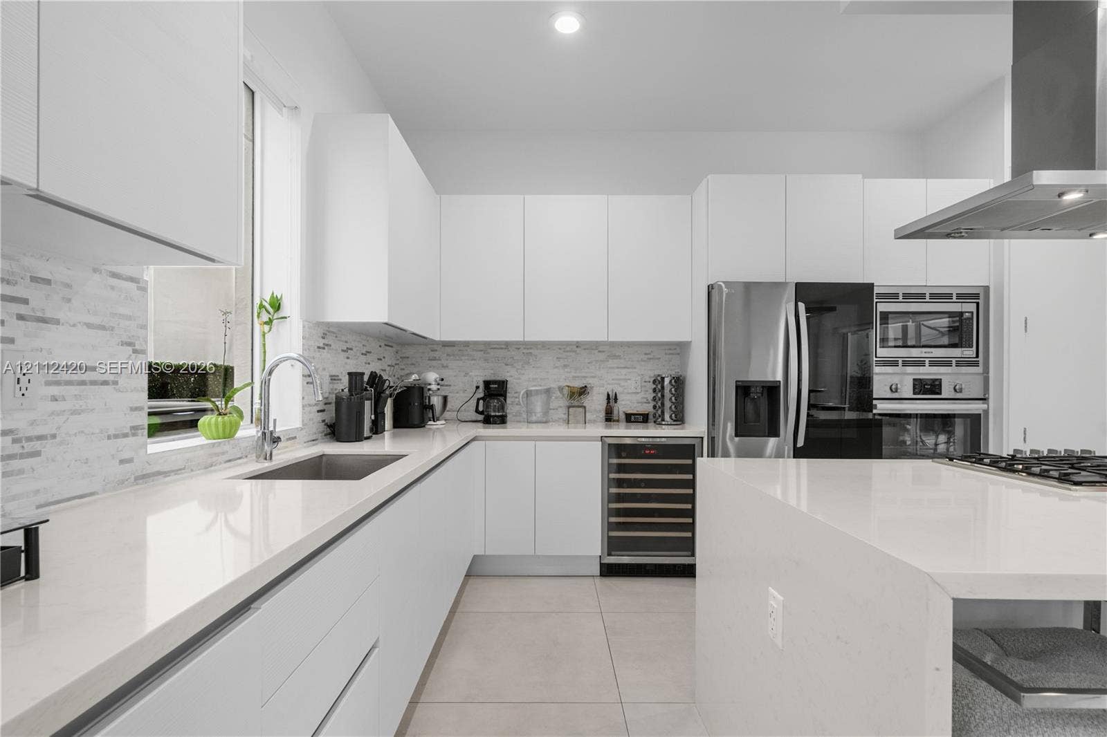 10054 NW 76th Terrace, Doral, FL 33178 Modern kitchen featuring white flat-panel cabinetry, quartz countertops, a tiled backsplash, stainless steel appliances including a built-in wine refrigerator, and a waterfall-edge kitchen island