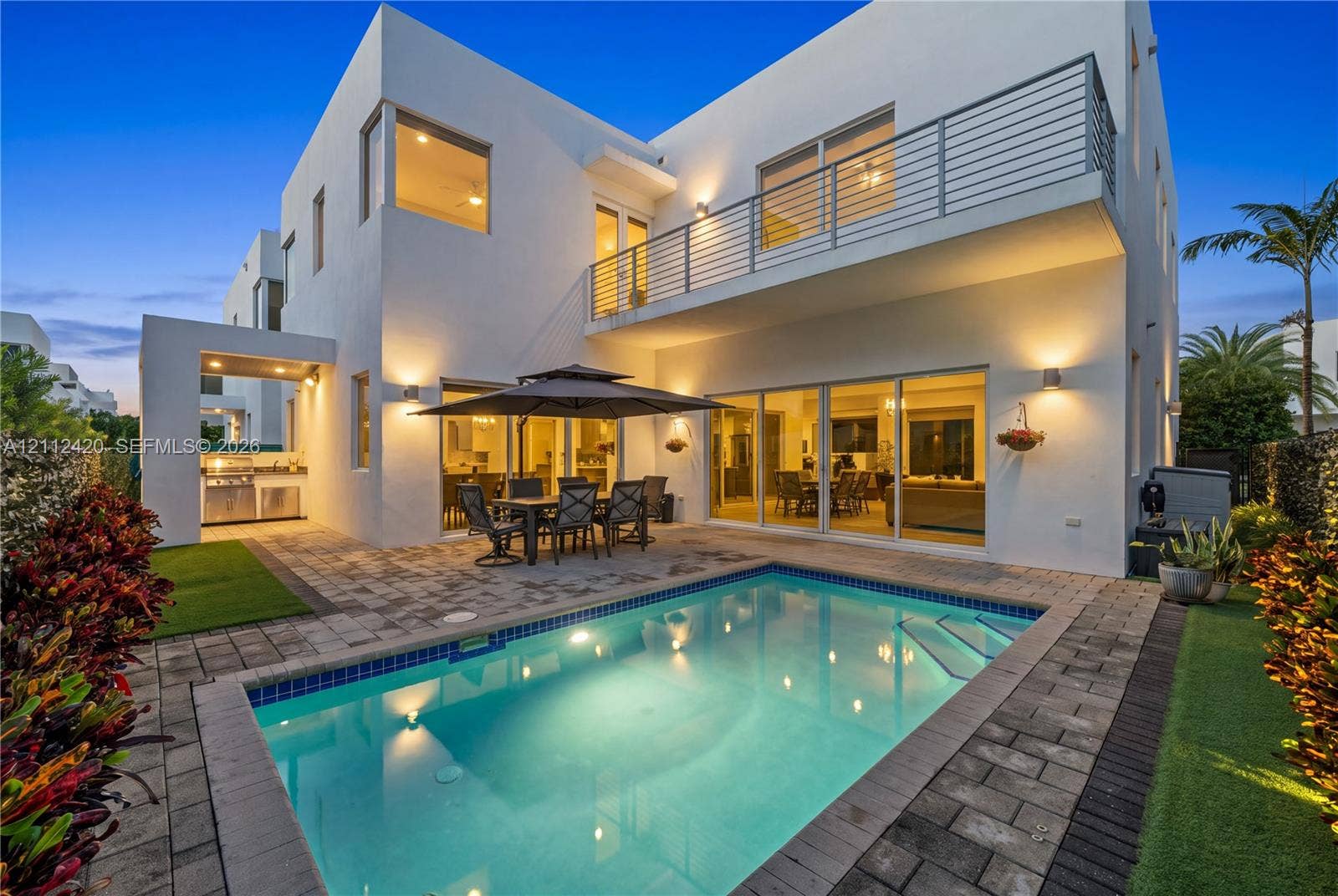 10054 NW 76th Terrace, Doral, FL 33178 Modern two-story residence featuring a paved pool deck, custom swimming pool, exterior kitchen with stainless steel grill, and an upper-level balcony with metal railing