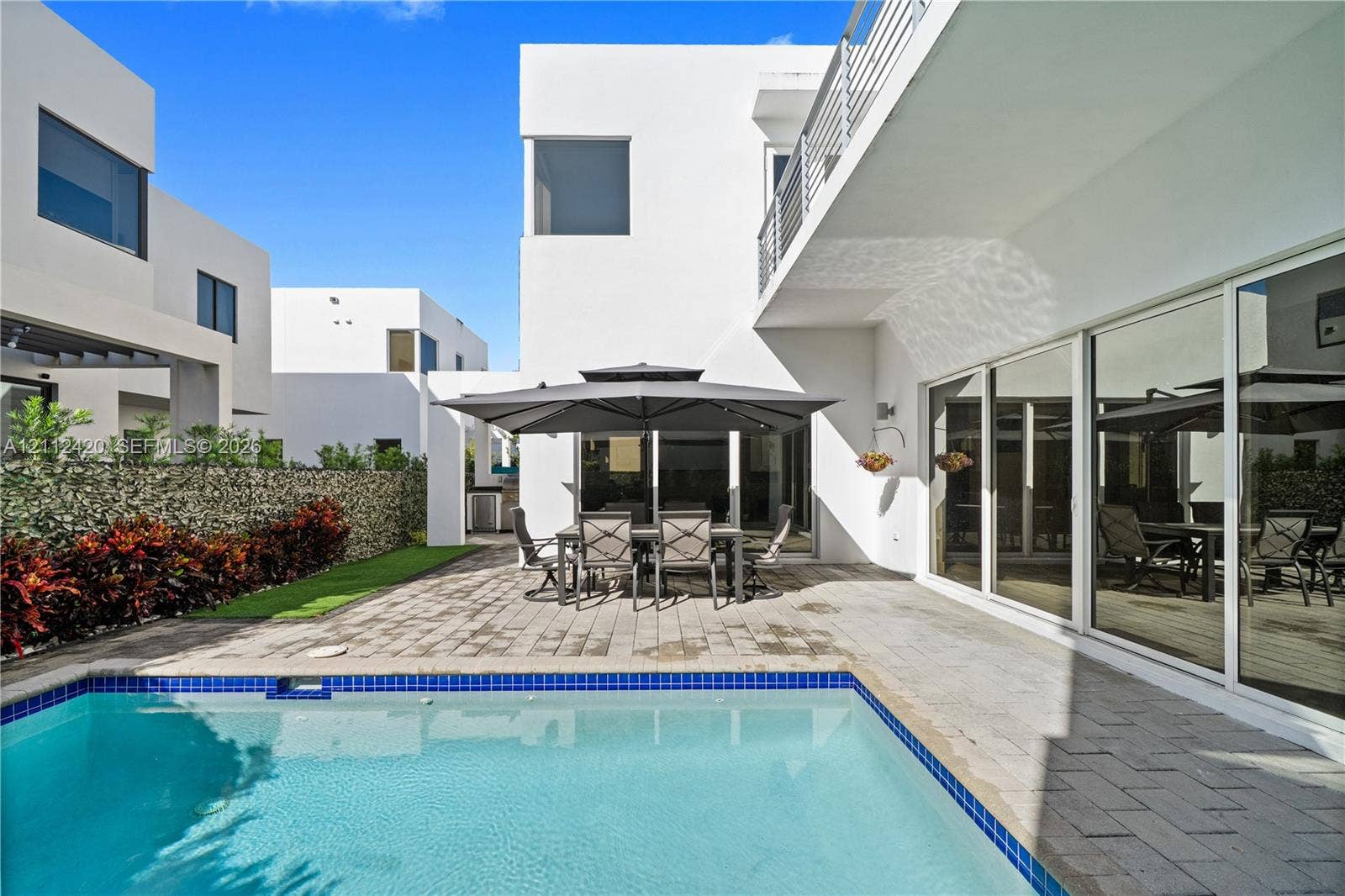10054 NW 76th Terrace, Doral, FL 33178 Modern stucco exterior with expansive sliding glass doors, paver patio, in-ground pool with blue tile border, and privacy hedge