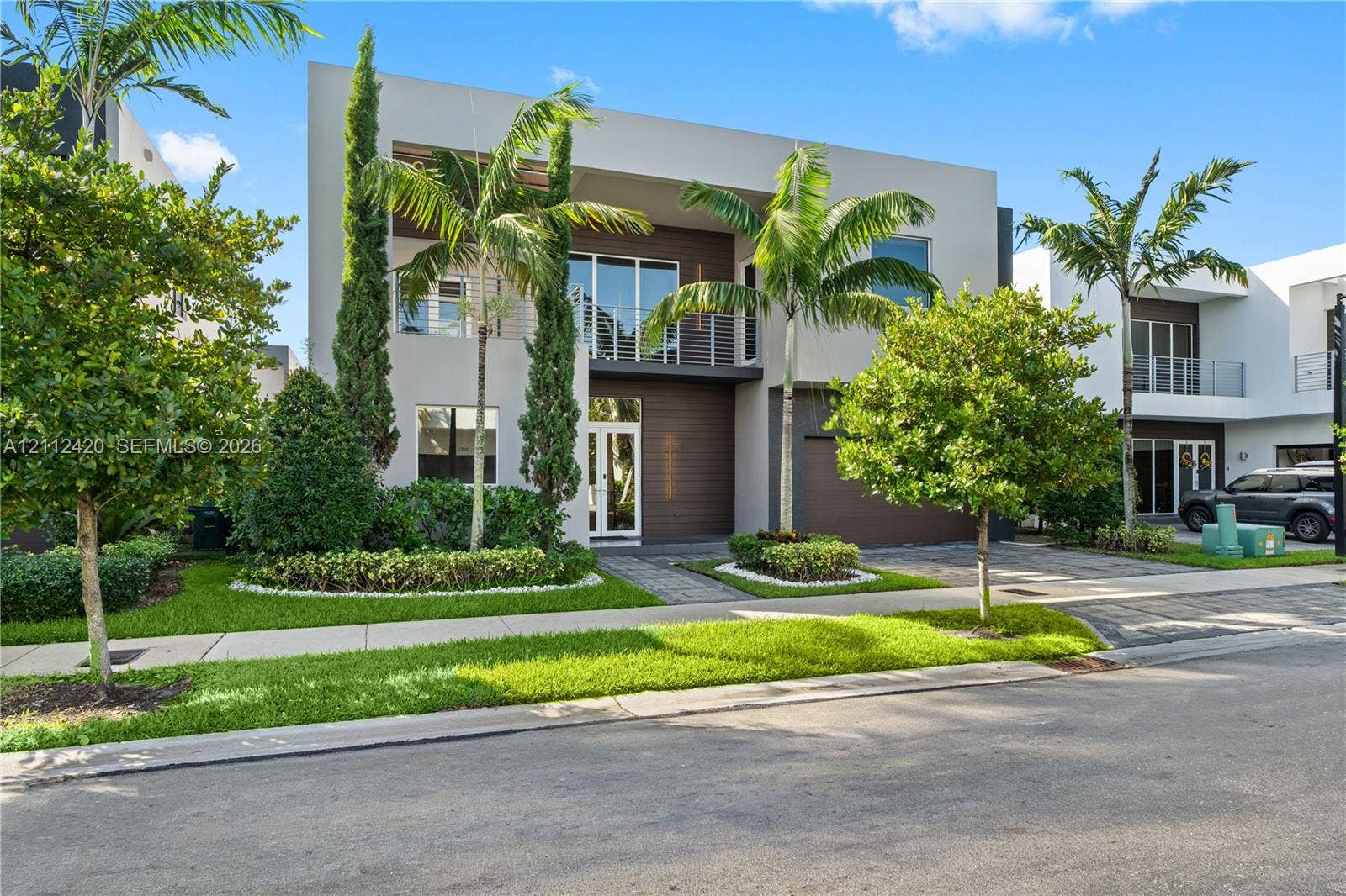 10054 NW 76th Terrace, Doral, FL 33178 Modern two-story residence featuring a smooth stucco finish, horizontal wood-tone siding, a glass-railed upper balcony, and paver driveway