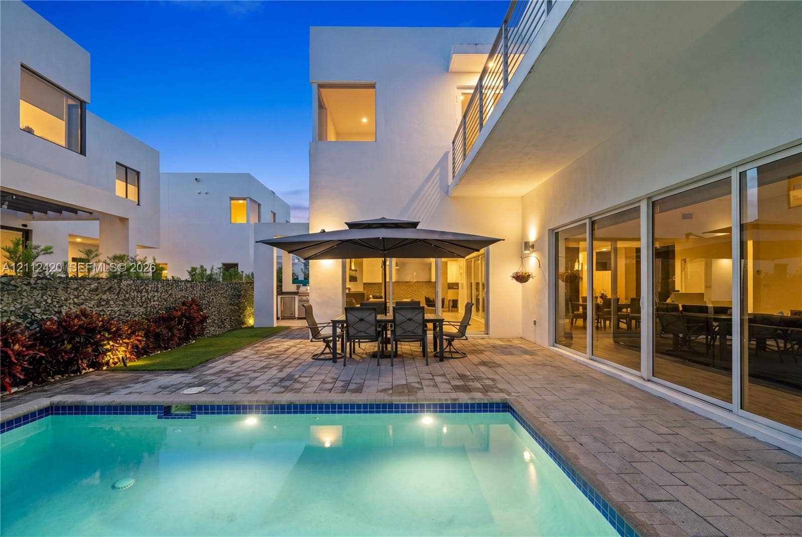 10054 NW 76th Terrace, Doral, FL 33178 Modern residence featuring a swimming pool with tile trim, expansive stone paver patio, minimalist stucco exterior, and multi-panel floor-to-ceiling glass sliding doors