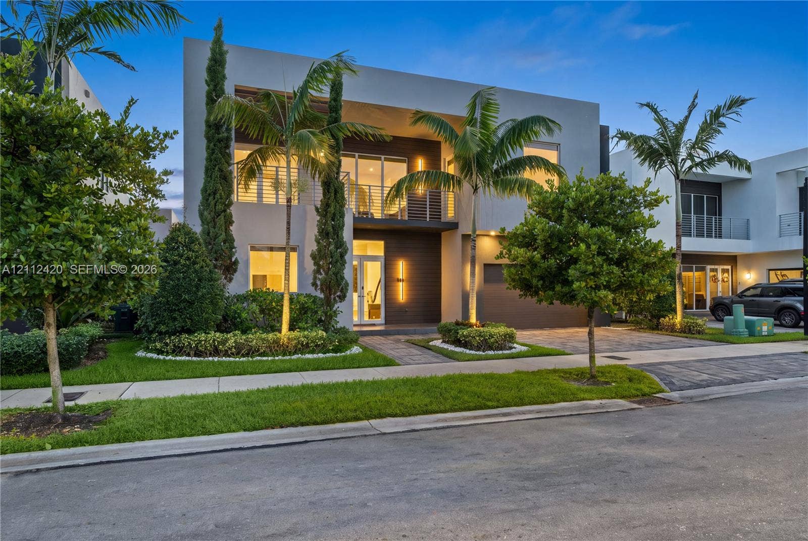 10054 NW 76th Terrace, Doral, FL 33178 Modern two-story exterior featuring a smooth stucco faade, horizontal wood-style cladding, expansive glass panels, a second-level balcony with metal railing, and paver driveway