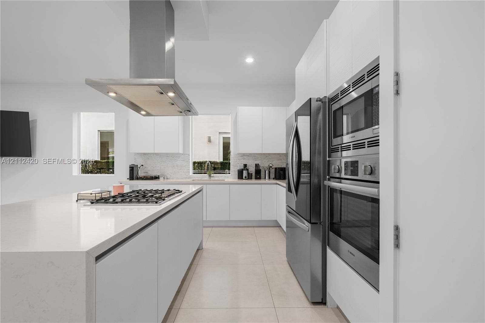 10054 NW 76th Terrace, Doral, FL 33178 Contemporary kitchen featuring handleless white cabinetry, solid-surface countertops, a stainless steel range hood, built-in stainless steel double ovens, and tile flooring