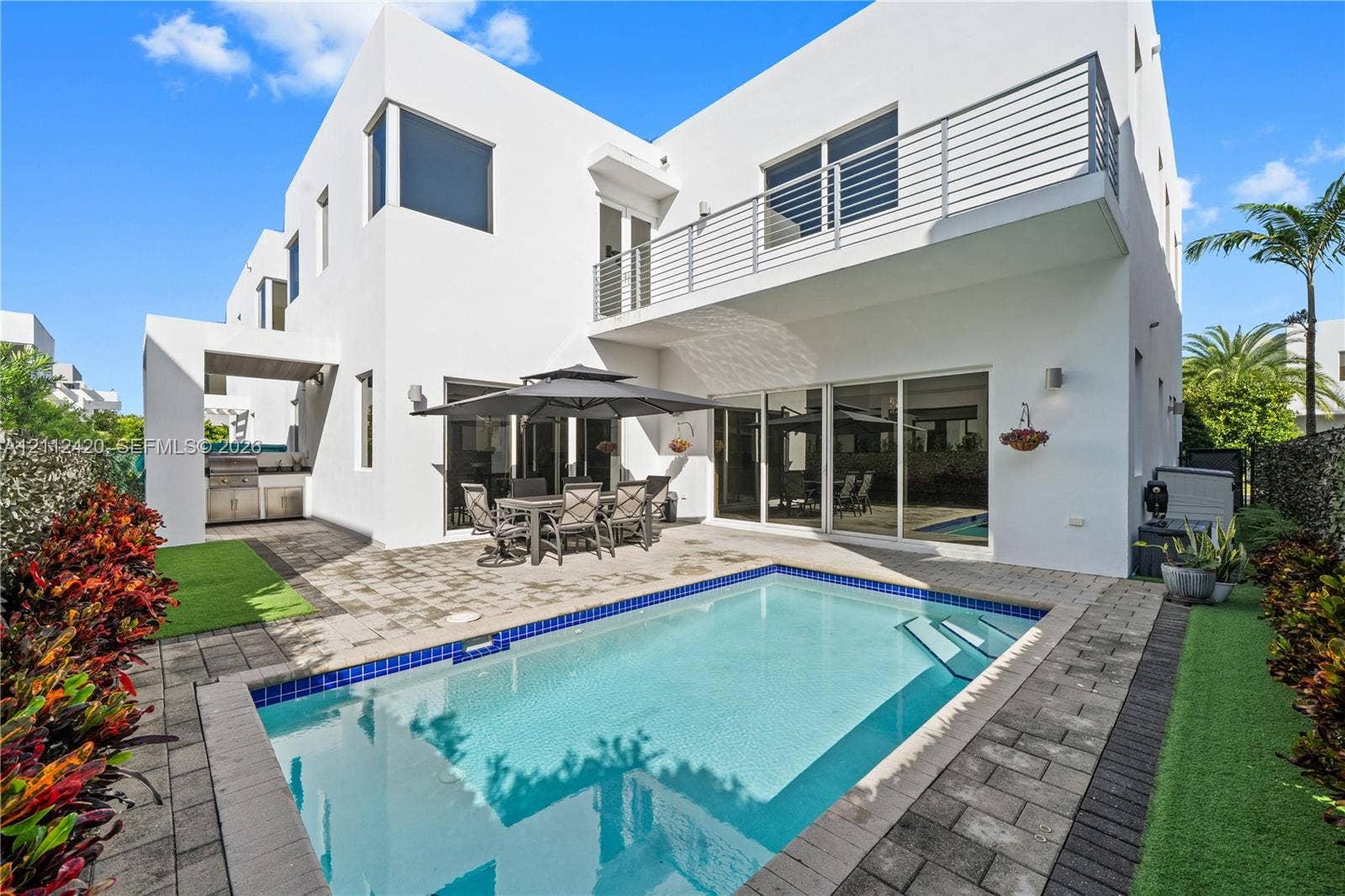 10054 NW 76th Terrace, Doral, FL 33178 Modern two-story stucco exterior featuring a built-in outdoor kitchen, second-level balcony with horizontal metal railings, concrete paver patio, and an in-ground swimming pool