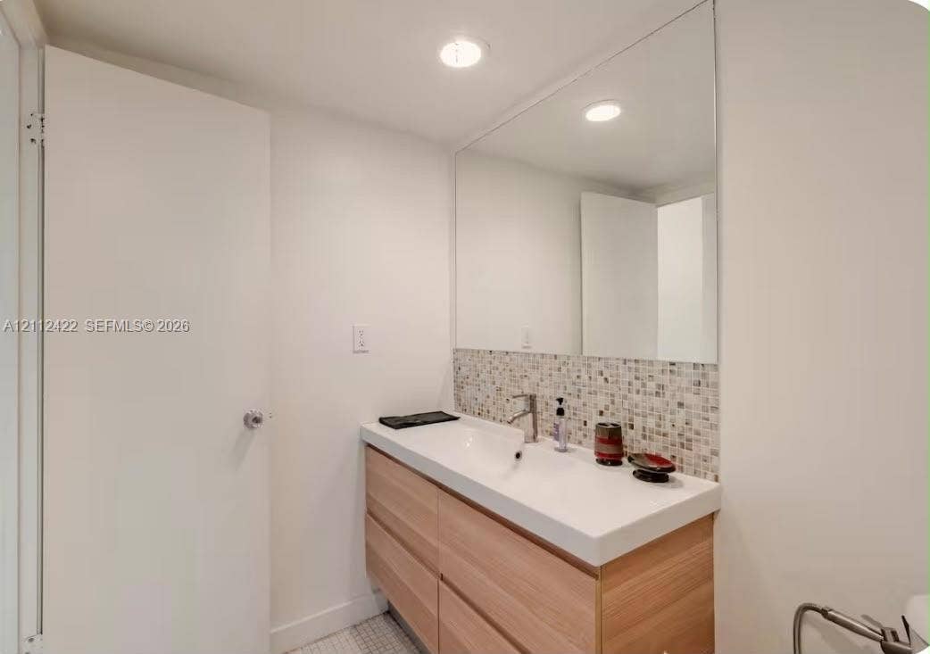 Floating wood-finish vanity with a rectangular sink, multi-toned mosaic tile backsplash, large wall-mounted mirror, and recessed ceiling lighting