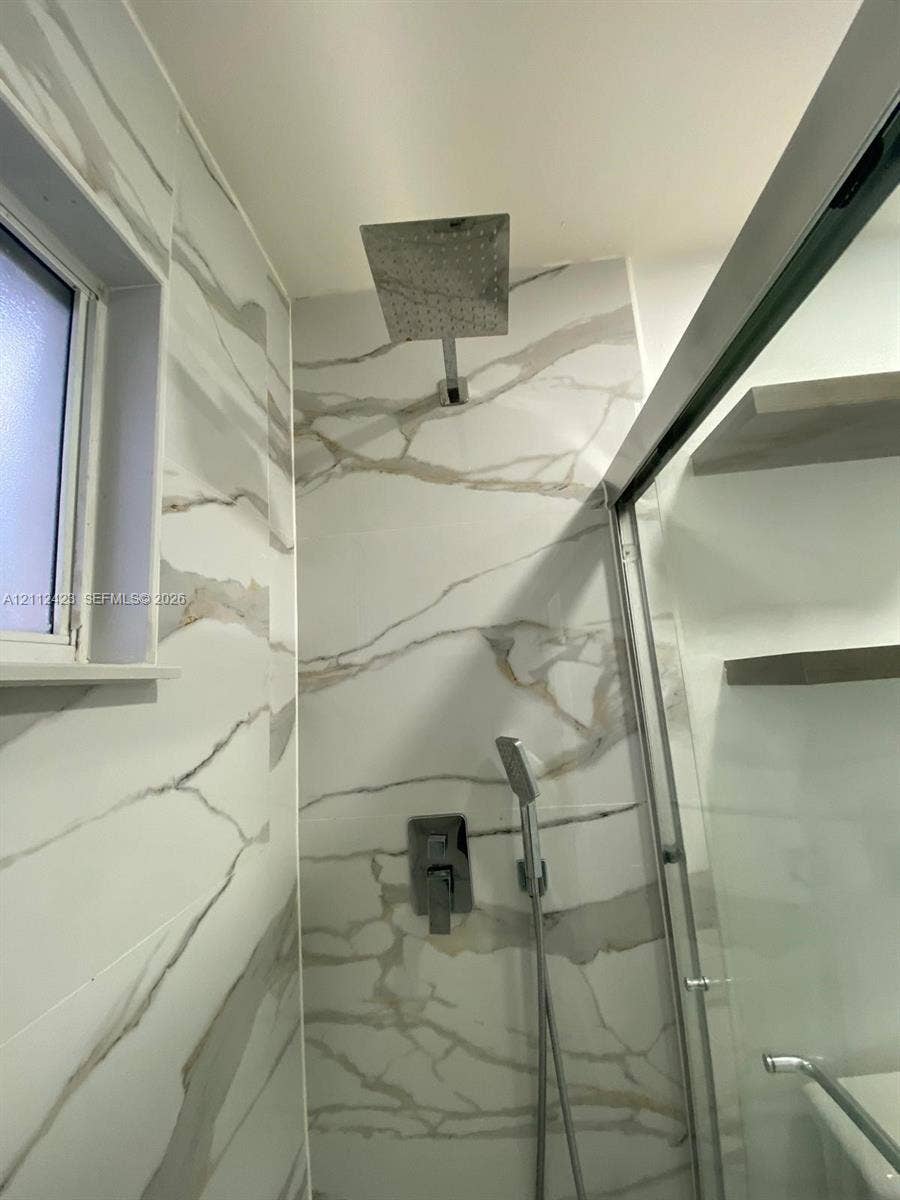 8555 SW 152nd Avenue, #104, Miami, FL 33193 Walk-in shower featuring large-format wall tile with veining, a rainfall showerhead, a separate handheld sprayer, and built-in wall shelving enclosed by a glass door