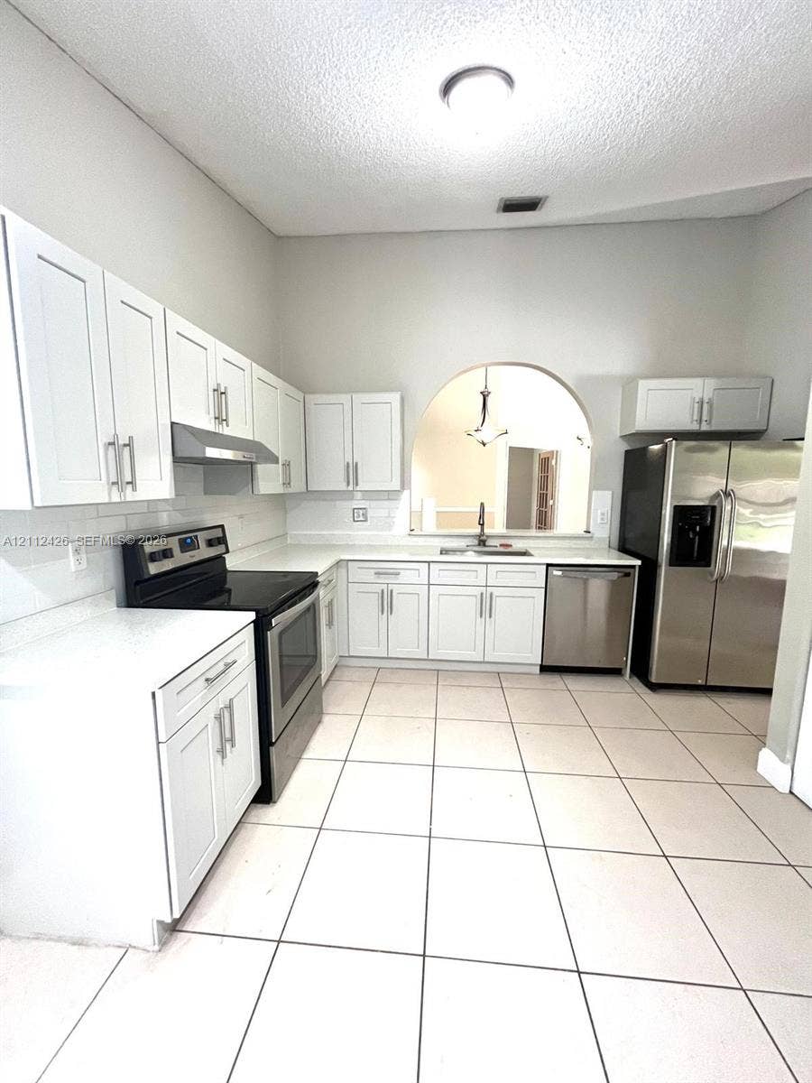 Kitchen featuring white shaker-style cabinetry, solid-surface countertops, subway tile backsplash, stainless steel appliances, and ceramic tile flooring with an arched pass-through window
