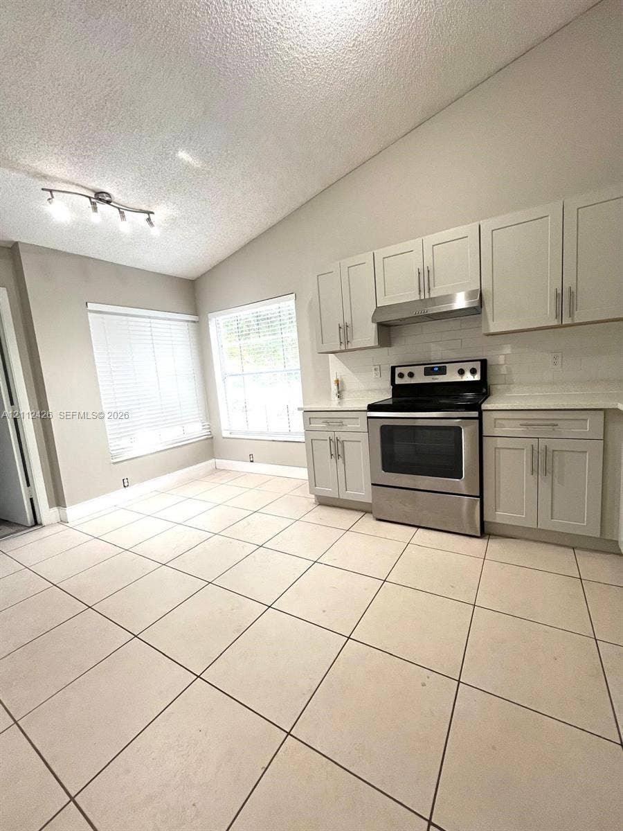 Kitchen featuring vaulted ceilings, light-tone cabinetry, stainless steel range with overhead vent hood, white tile backsplash, and oversized floor tiles