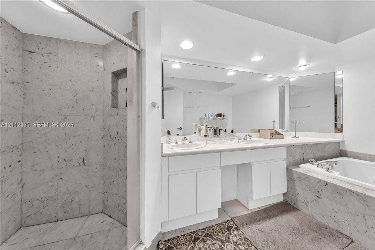 Bathroom featuring stone tile shower walls with an integrated recessed niche, dual-sink vanity with white cabinetry, and a stone-surround soaking tub