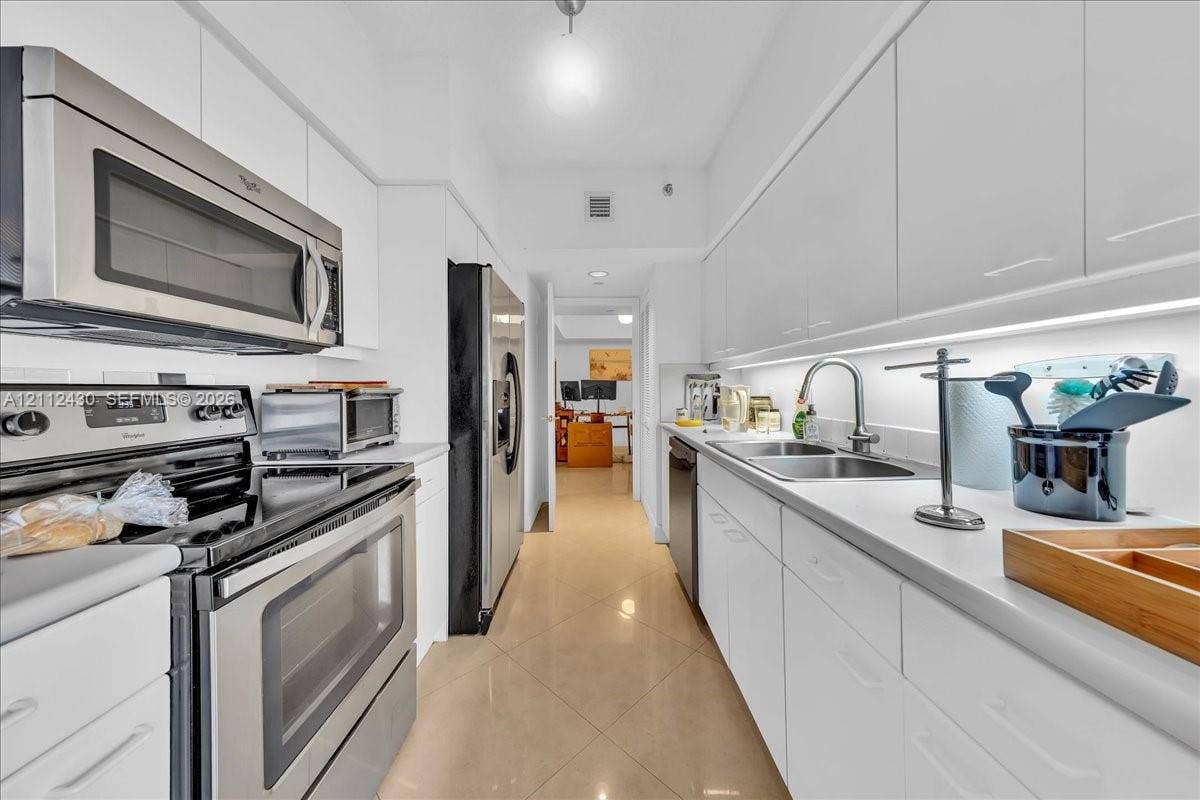 Galley kitchen featuring high-gloss white cabinetry, stainless steel appliances, solid-surface countertops, dual-basin sink with arched faucet, and ceramic tile flooring