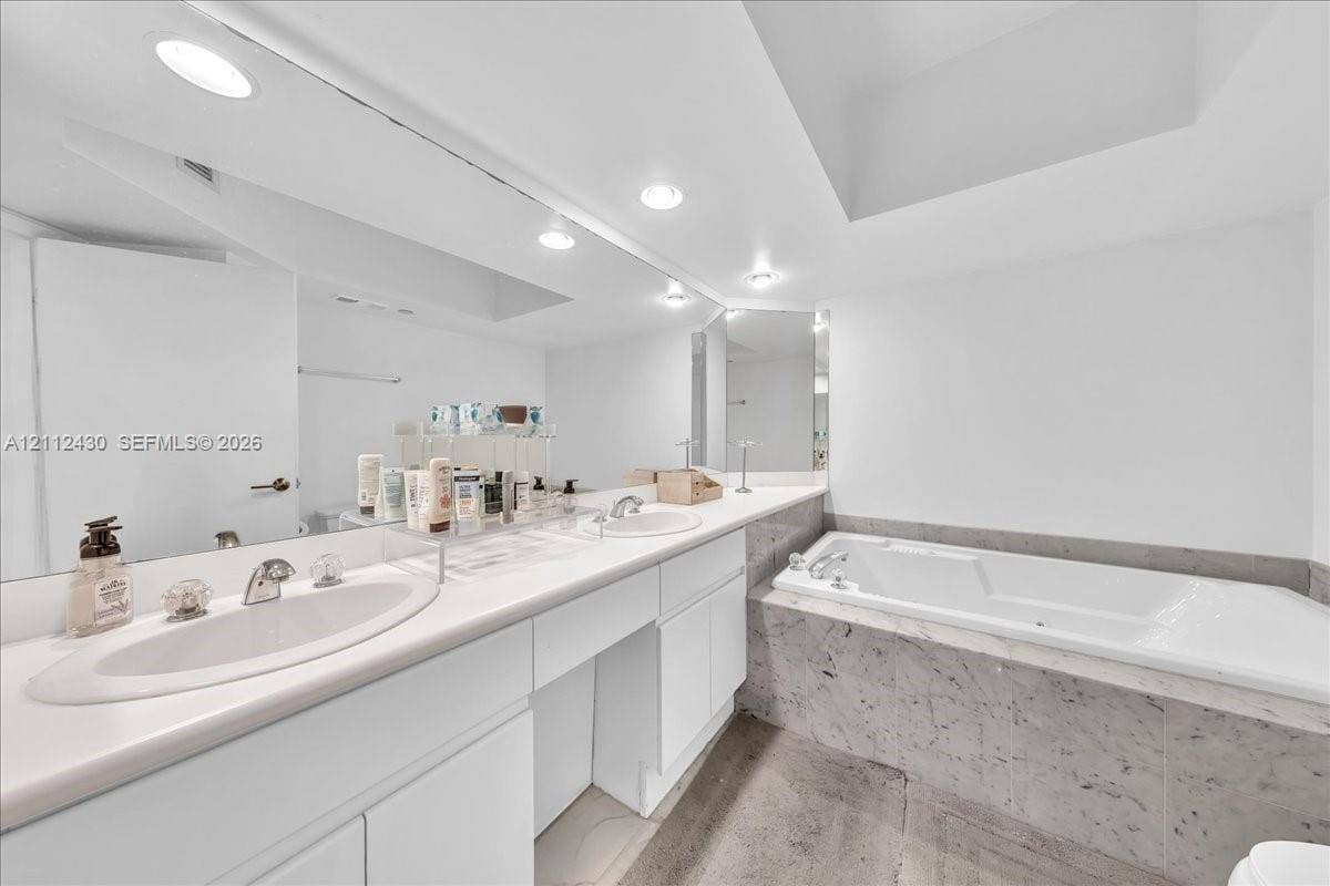 Dual-sink vanity with white cabinetry and solid surface countertop, large wall-to-wall mirror, recessed lighting, and a drop-in soaking tub with stone-finish surround