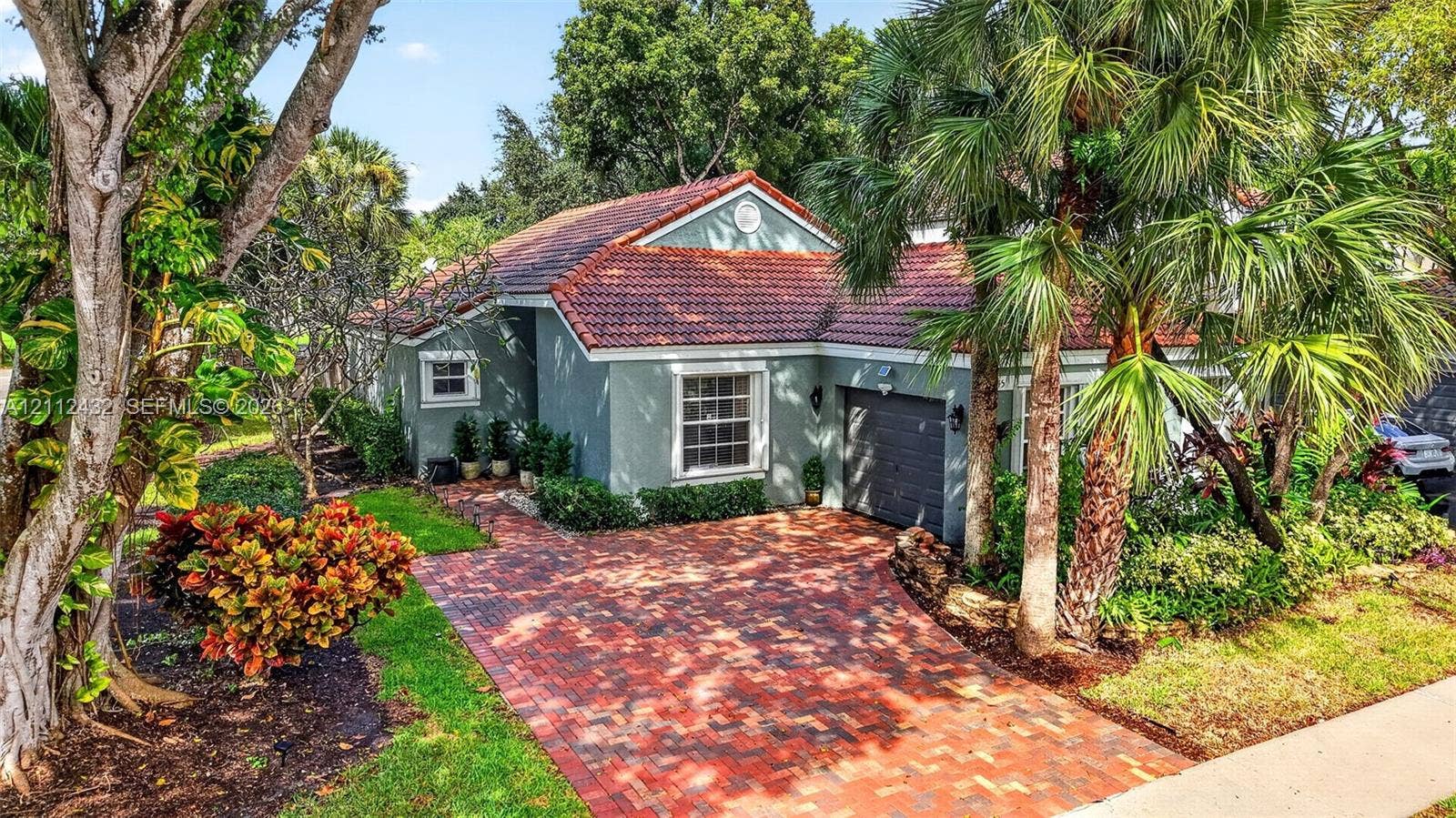 10545 NW 11th Court, Plantation, FL 33322 Single-story stucco exterior featuring a red tile roof, an expansive brick-paver driveway, and mature tropical landscaping