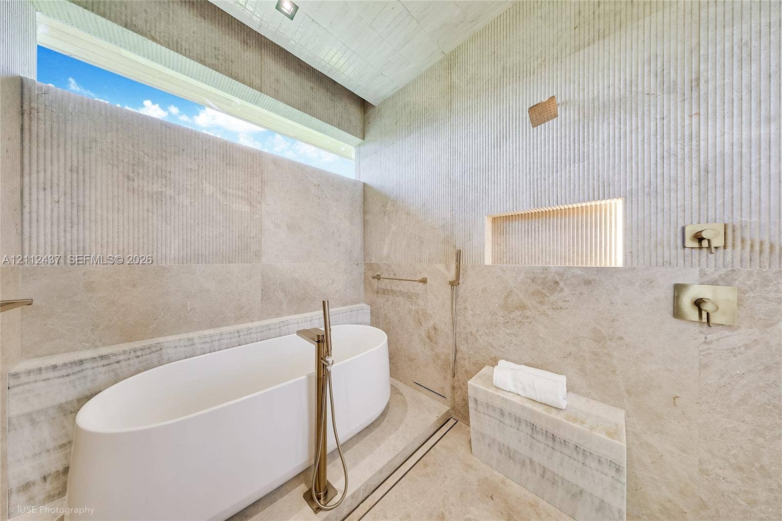 770 Lake Road, Miami, FL 33137 Freestanding white soaking tub with a floor-mounted faucet, floor-to-ceiling stone wall tiling, integrated recessed niche with accent lighting, built-in stone bench, and horizontal clerestory window