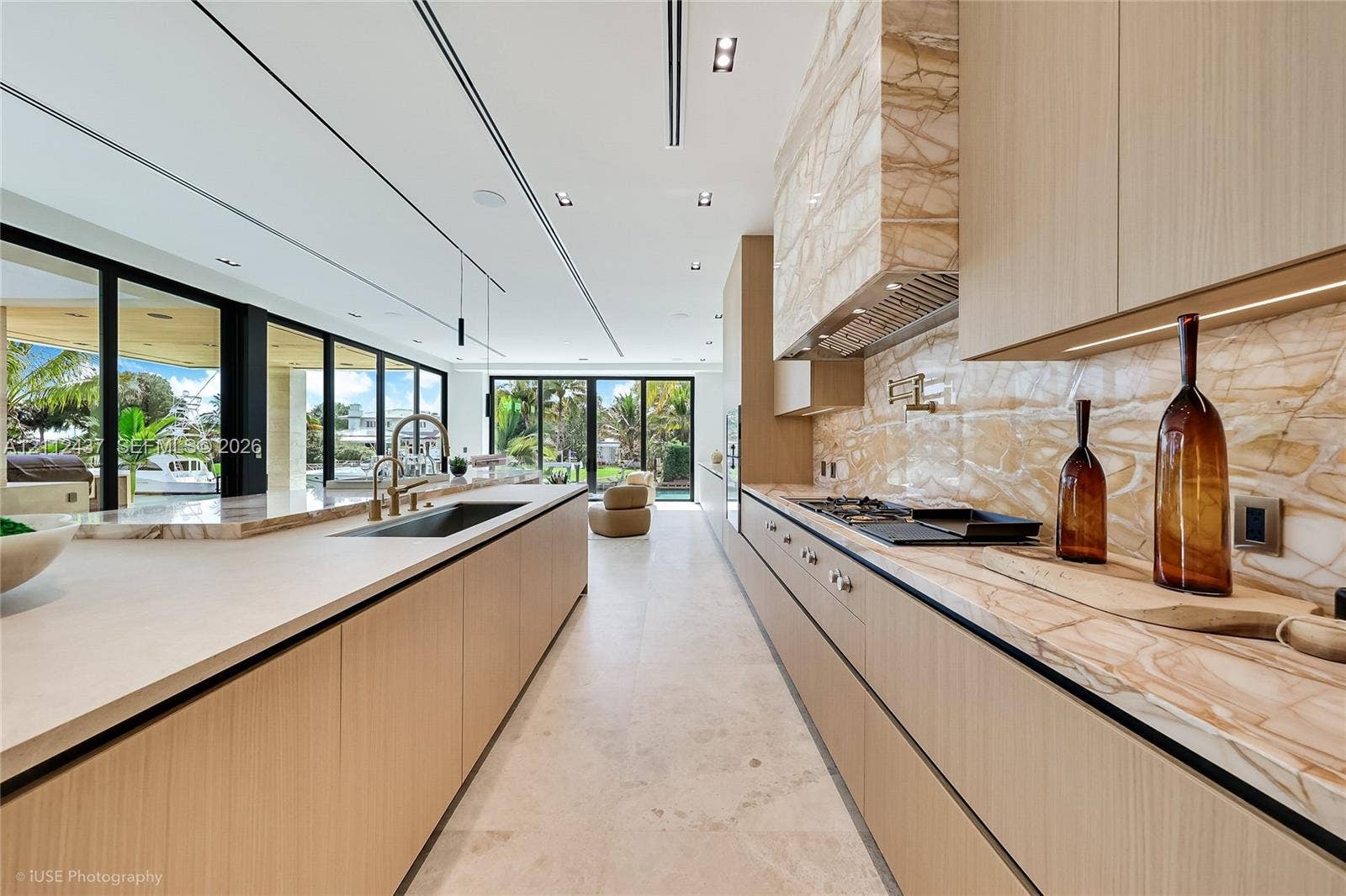 770 Lake Road, Miami, FL 33137 Custom wood-finish cabinetry, expansive stone-topped kitchen island with waterfall edge, integrated stainless steel appliances, and floor-to-ceiling glass sliding doors