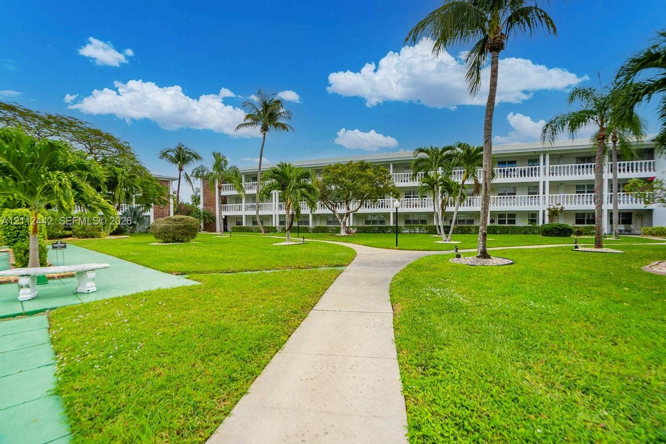 2455 NE 51st Street, #E114, Fort Lauderdale, FL 33308 Mid-rise multi-story building exterior featuring open-air corridors with decorative balustrade railings, manicured green lawns, paved walkways, and mature palm trees