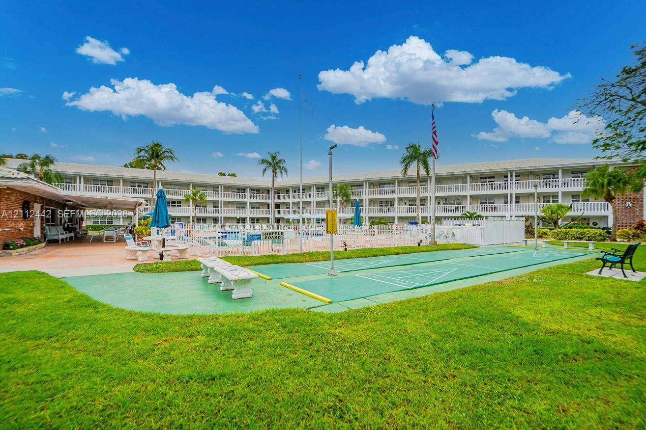 2455 NE 51st Street, #E114, Fort Lauderdale, FL 33308 Outdoor recreation area featuring a shuffleboard court, a swimming pool with enclosure, a landscaped lawn, and palm trees