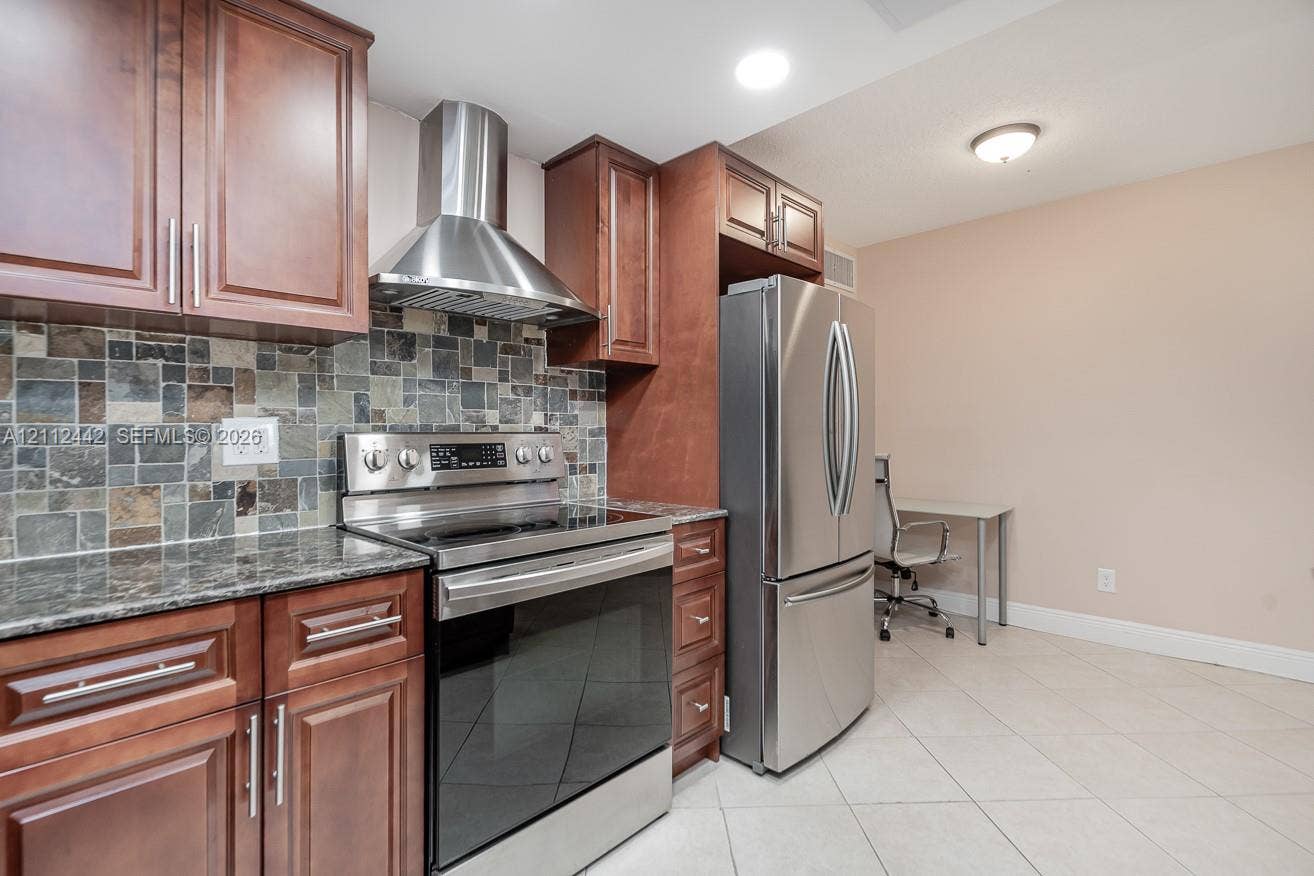 2455 NE 51st Street, #E114, Fort Lauderdale, FL 33308 Kitchen featuring stained wood cabinetry, stone-tiled backsplash, polished stone countertops, stainless steel appliances, and tile flooring