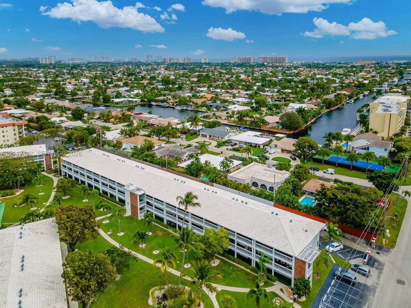 2455 NE 51st Street, #E114, Fort Lauderdale, FL 33308 Aerial perspective featuring a multi-story residential building with a light-colored pitched roof, surrounded by landscaped grounds with mature palm trees, paved walkways, and dedicated surface parking areas