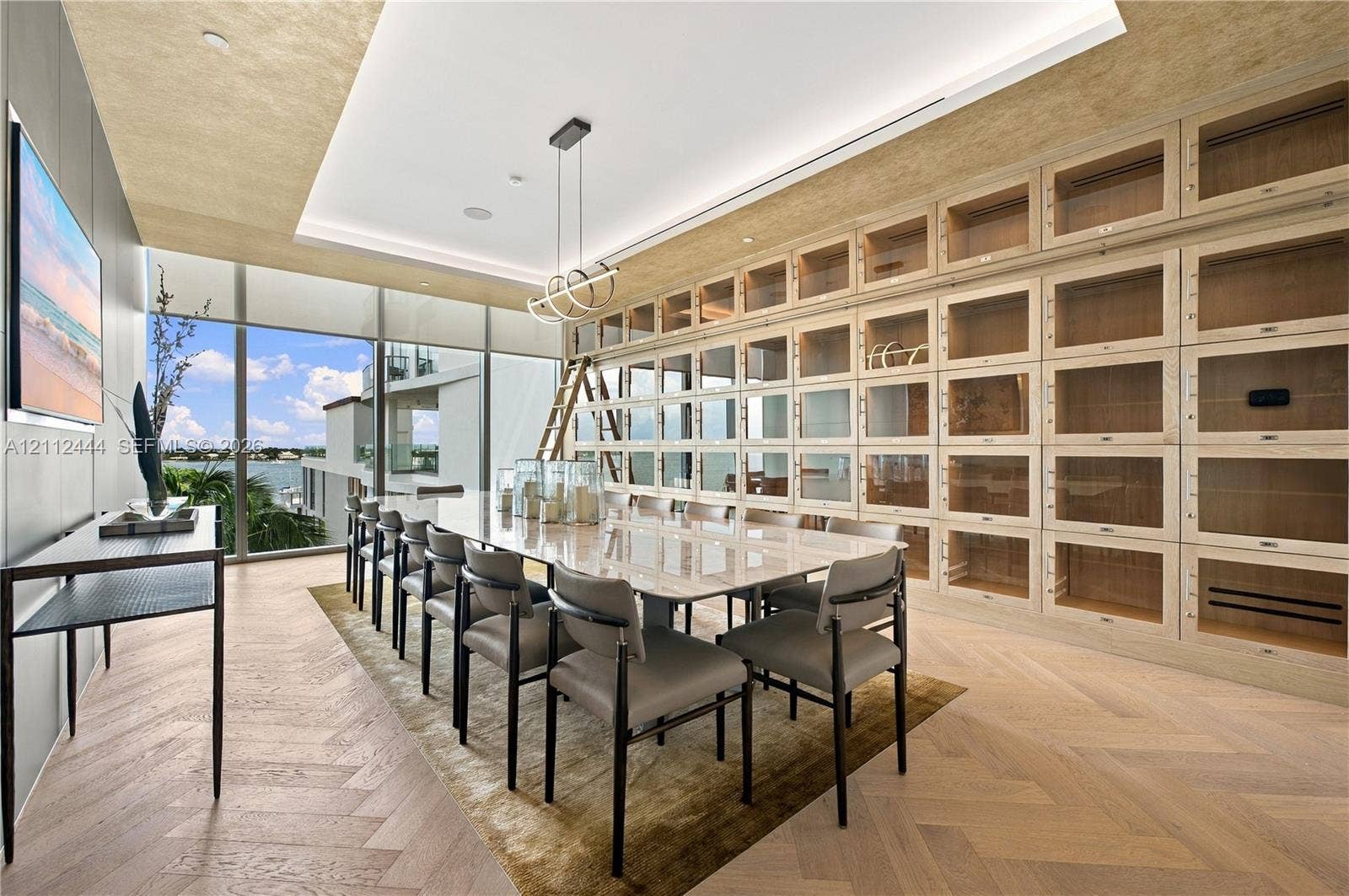 Floor-to-ceiling built-in cabinetry with integrated rolling library ladder, wood-finish herringbone flooring, recessed tray ceiling, modern linear pendant lighting, and full-height glass windows