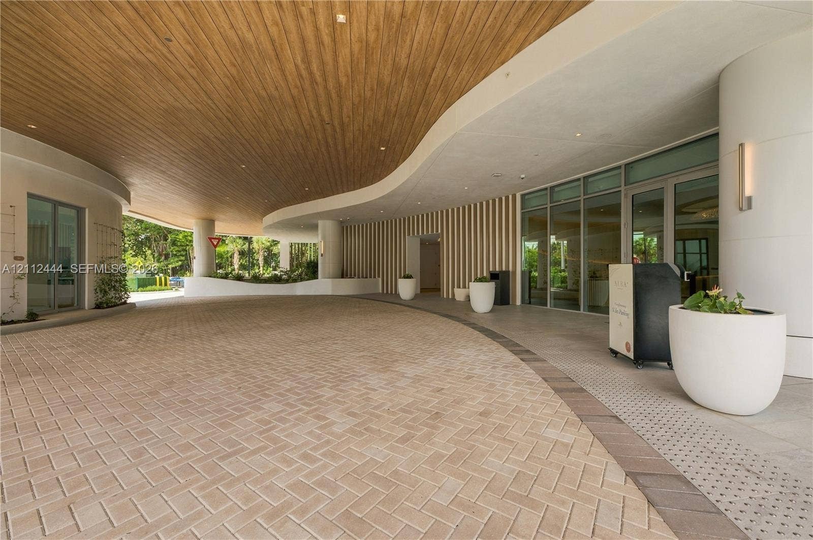 Covered porte-cochre featuring a curved wood-panel ceiling, herringbone-pattern paver drive, vertical slat wall detailing, and expansive glass entry doors