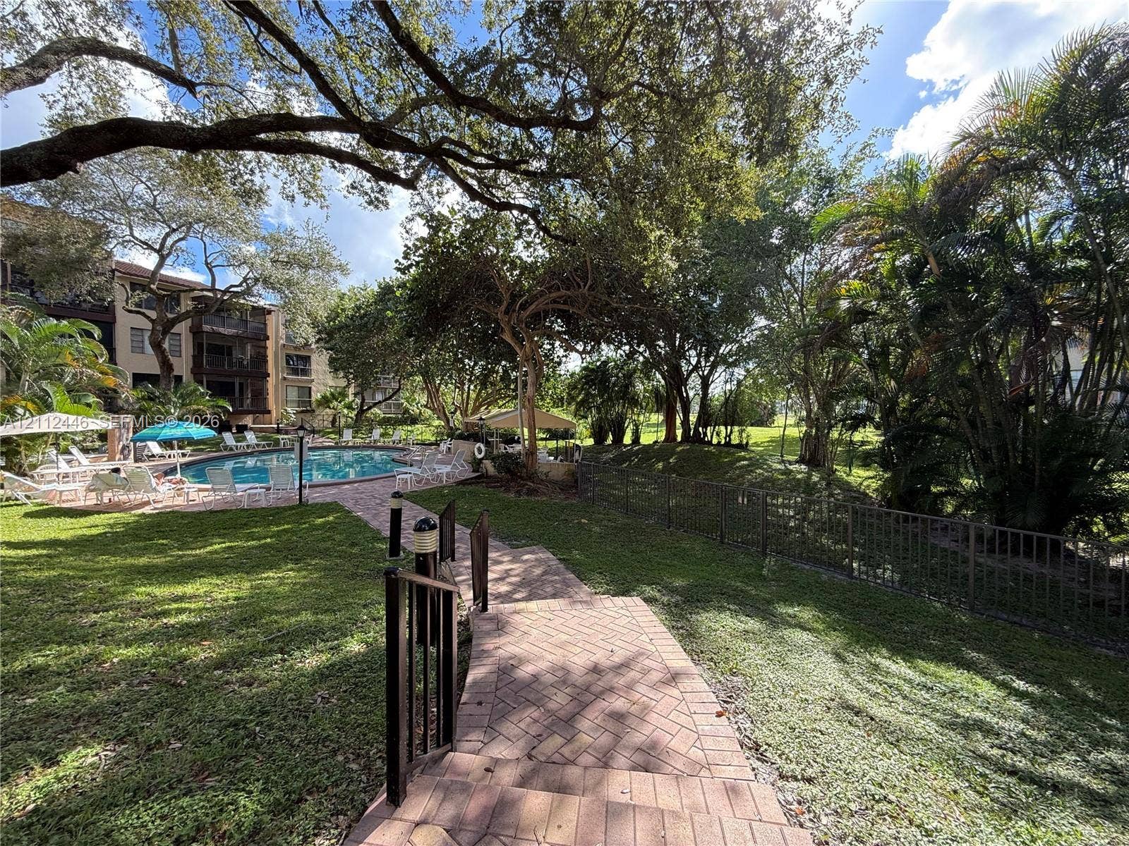 6100 S Falls Cir Drive, #411, Lauderhill, FL 33319 Outdoor pool with brick-paver deck, landscaped grounds, mature shade trees, and metal perimeter fencing