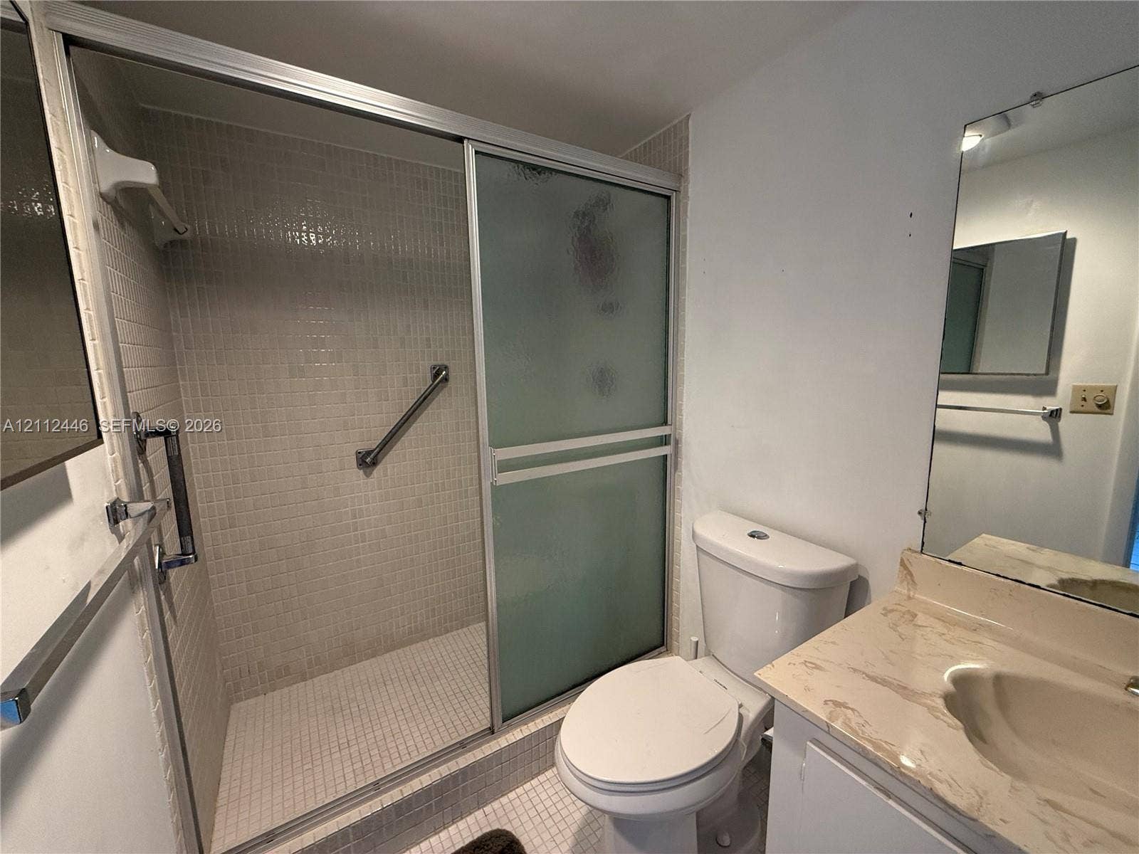 6100 S Falls Cir Drive, #411, Lauderhill, FL 33319 Bathroom featuring a walk-in shower with frosted sliding glass doors, mosaic tile surround, and mounted grab bars, paired with a vanity with a stone-finish countertop and integrated sink