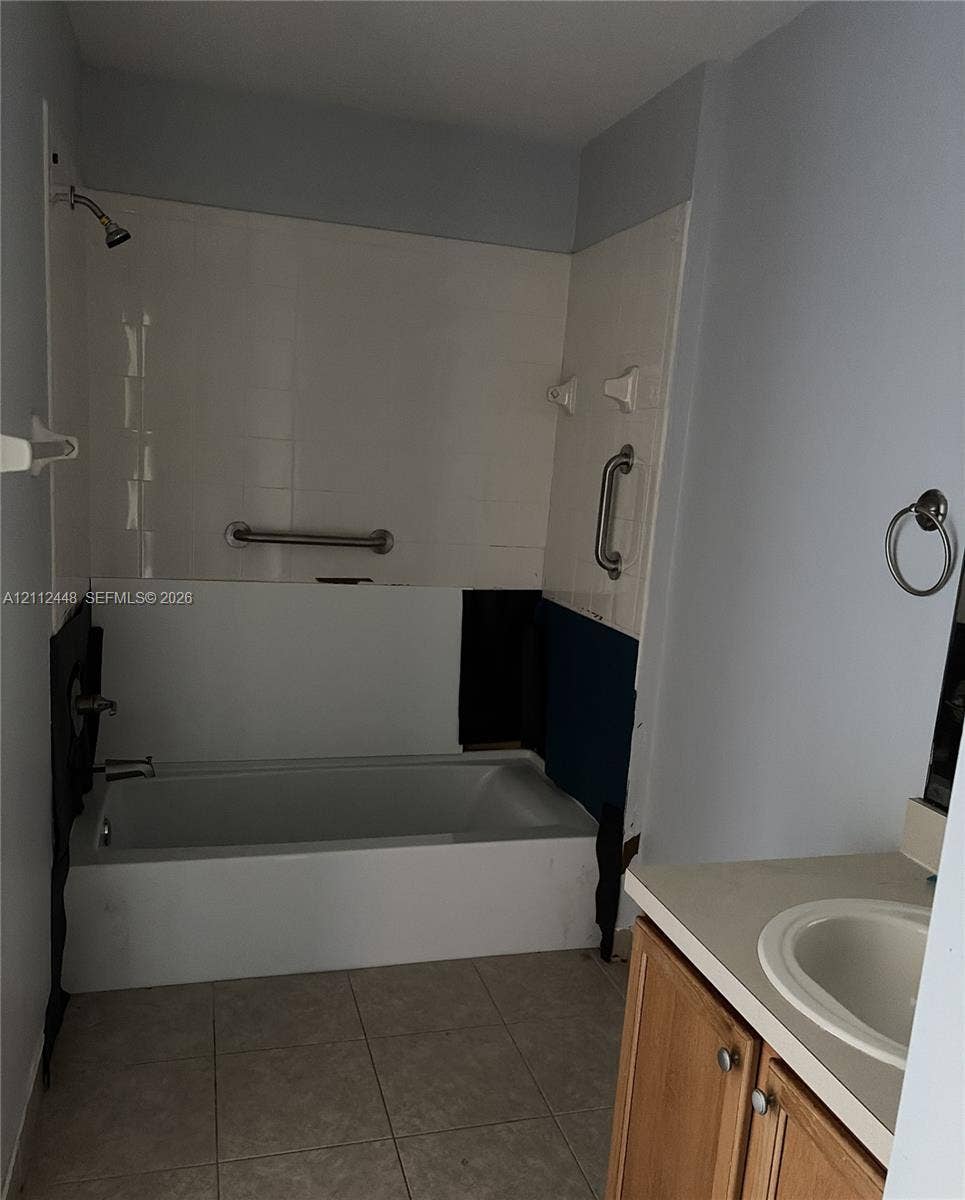 2007 SW Marblehead Way, Port St Lucie, FL 34953 Bathtub with wall-mounted shower head, tile surround, and installed safety grab bars, paired with a wood-finish vanity featuring an integrated basin and a neutral countertop